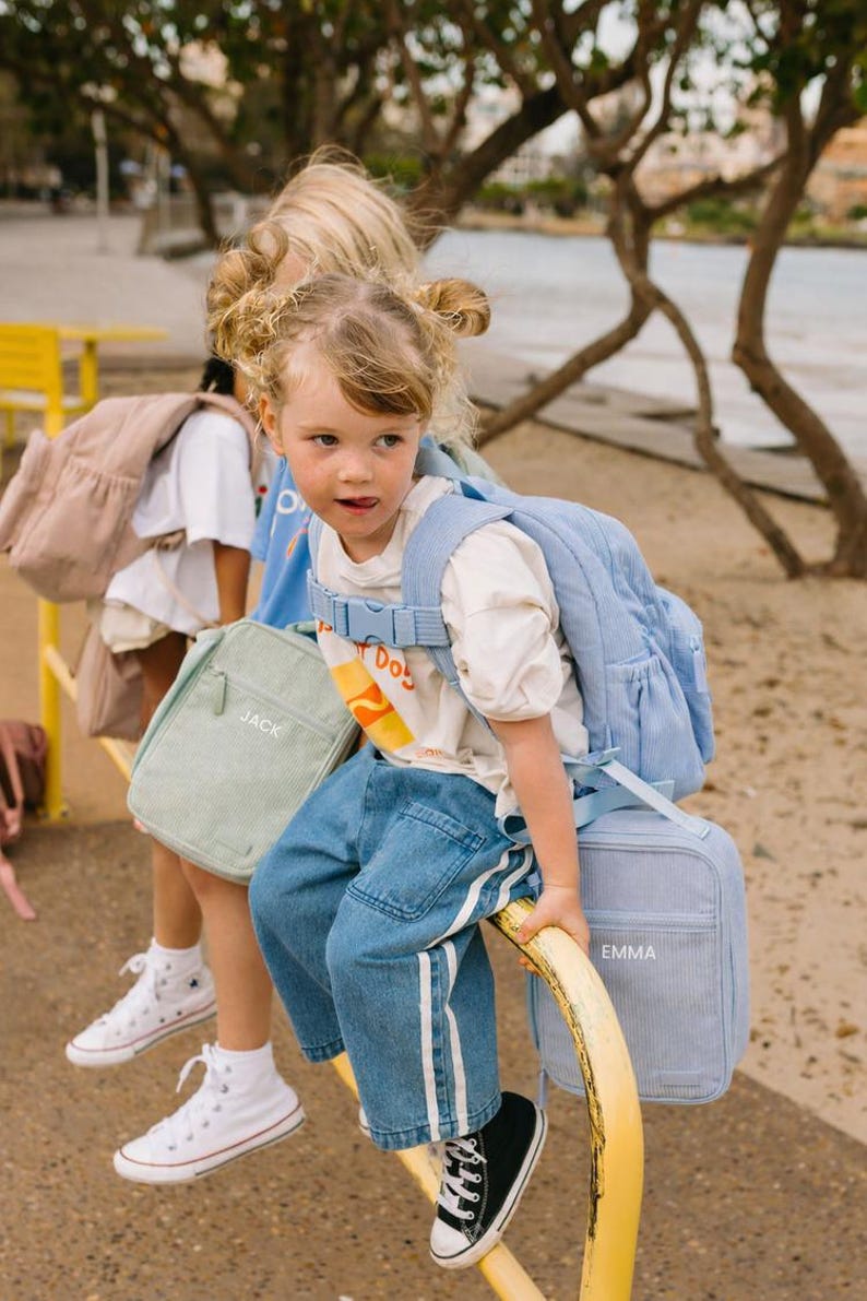 May include: Two young children with backpacks and lunch bags. One child wears a light blue backpack and matching lunch bag with the name "Emma". The other child has a light green lunch bag with the name "Jack". Both children are wearing sneakers.
