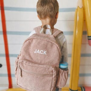 May include: A pink corduroy backpack with the name "JACK" embroidered in white. The backpack has a front zippered pocket and a side pocket holding a blue water bottle. The child is wearing the backpack.