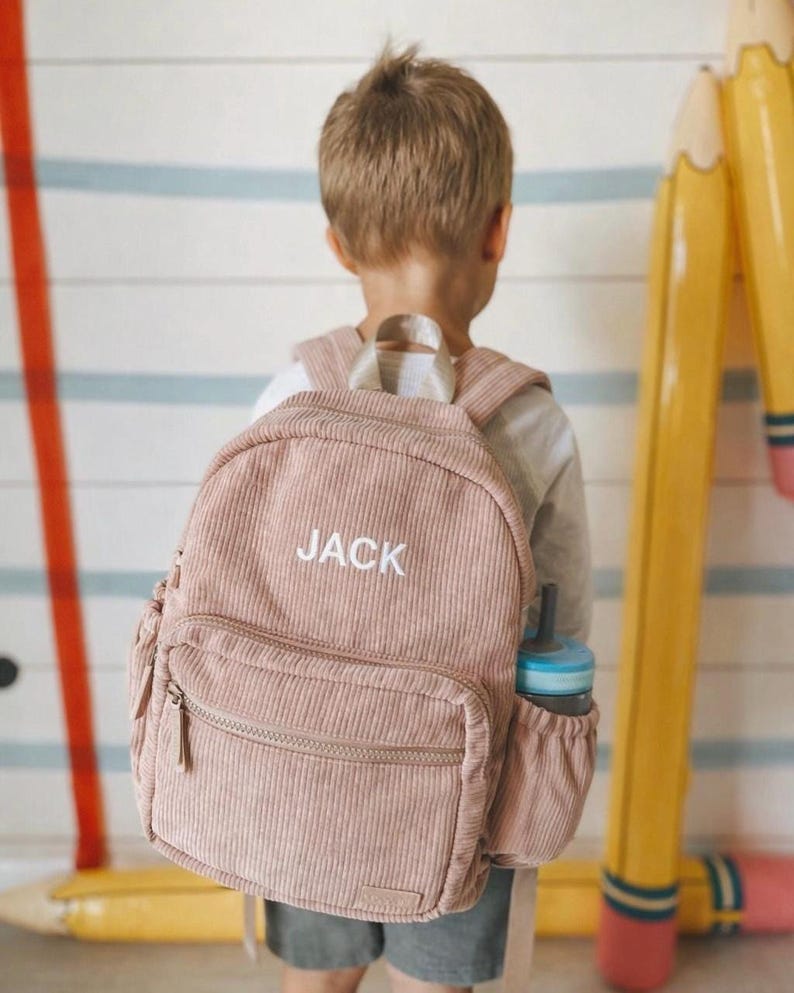 May include: A pink corduroy backpack with the name "JACK" embroidered in white. The backpack has a front zippered pocket and a side pocket holding a blue water bottle. The child is wearing the backpack.
