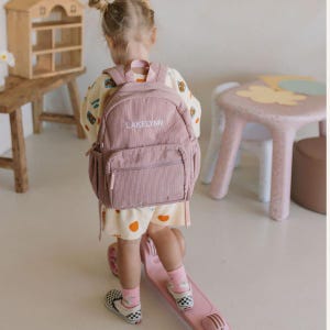 May include: A dusty rose corduroy backpack with the name "LAKELYNN" embroidered in white. The backpack has a front pocket and side pockets. The child is wearing a patterned shirt and shorts, pink socks, and checkered shoes.