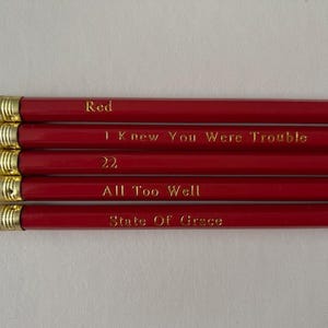 Op de afbeelding: Vijf rode potloden met goudkleurige metalen accenten en witte gummen. Elk potlood heeft een andere zin in goud gedrukt: "Red", "I Knew You Were Trouble", "22", "All Too Well" en "State of Grace".