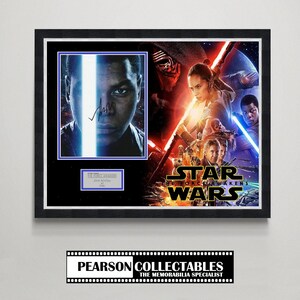 May include: Framed Star Wars: The Force Awakens film poster with John Boyega's autograph. The poster is in a black frame and features characters from the movie, with the title "Star Wars: The Force Awakens" clearly visible.