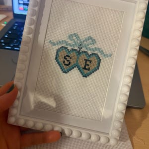 May include: White framed cross-stitch artwork showing two teal hearts with the letters "S" and "E". A teal bow joins the hearts. The frame has a decorative, rounded edge.
