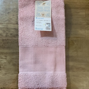 May include: A pink kitchen towel with a fringed border. The towel is made from 100% cotton and is part of the Kitchen Mates collection. The label reads "Cross Stitch Collectibles" and "Kitchen Mates/TT 11-6103-5 Pink Souffle 100% Cotton".