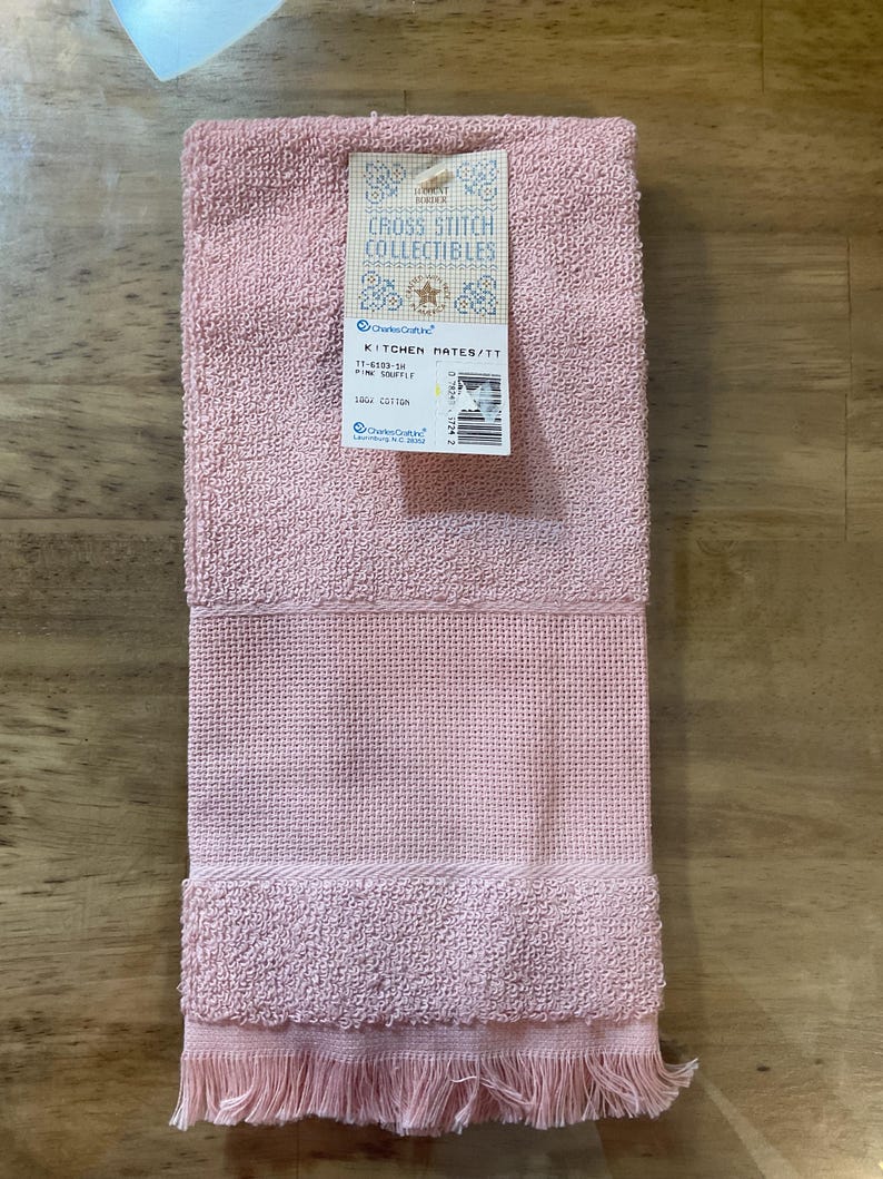 May include: A pink kitchen towel with a fringed border. The towel is made from 100% cotton and is part of the Kitchen Mates collection. The label reads "Cross Stitch Collectibles" and "Kitchen Mates/TT 11-6103-5 Pink Souffle 100% Cotton".