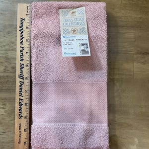 May include: A pink kitchen towel with a fringe border. The towel is made from 100% cotton and has the text "Kitchen Mates/TT" and "Cross Stitch Collectibles" on the label.