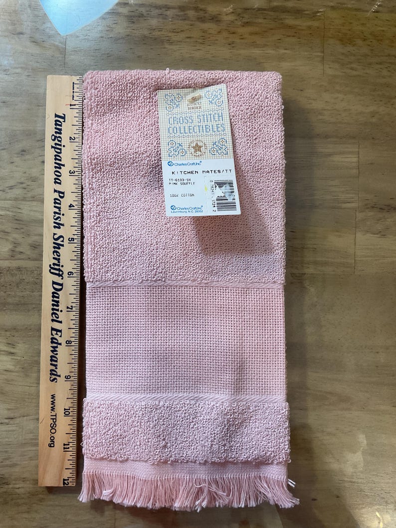 May include: A pink kitchen towel with a fringe border. The towel is made from 100% cotton and has the text "Kitchen Mates/TT" and "Cross Stitch Collectibles" on the label.