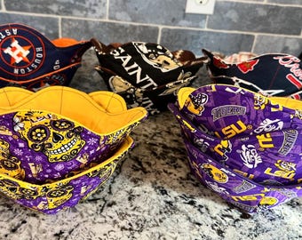 May include: A collection of fabric bowl cozies in various patterns and colors. Designs include a sugar skull, team logos, and other graphics. The cozies have a scalloped edge and are lined with a contrasting color.