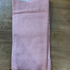 May include: A pink kitchen towel with a fringed edge. The towel is folded in half and has a white plastic tag attached.