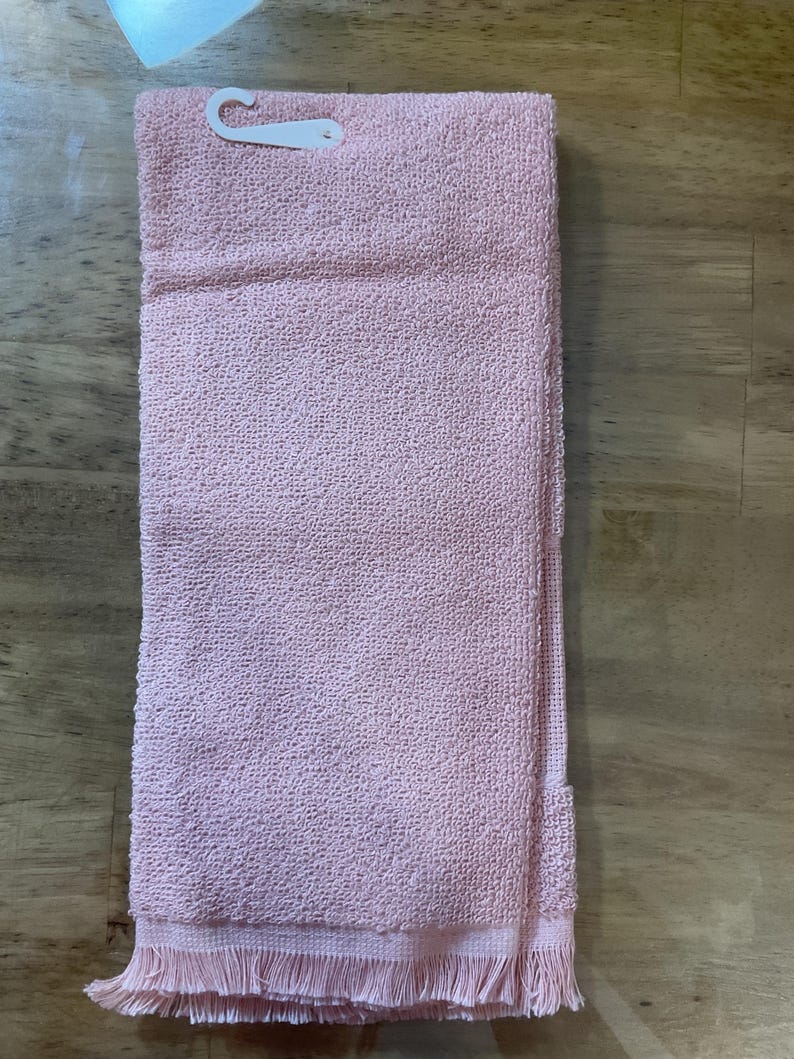 May include: A pink kitchen towel with a fringed edge. The towel is folded in half and has a white plastic tag attached.