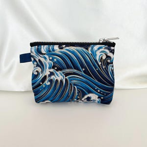 May include: A small, rectangular pouch with a zipper closure. The pouch features a blue and white wave pattern with gold accents. A small blue tab is attached to the side. The zipper is silver and the trim is black.