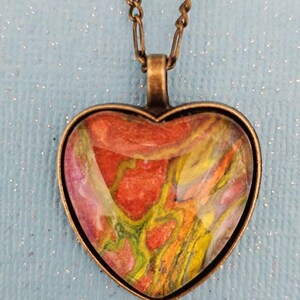 May include: A heart-shaped pendant necklace with a colorful abstract design inside the glass. The pendant is made of bronze metal and hangs from a bronze chain.