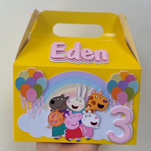 Pode incluir: Uma caixa de lembrancinhas amarela com uma alça, decorada com personagens de desenhos animados, um arco-íris, balões e o número "3". O nome "Eden" é exibido em letras rosa na caixa.