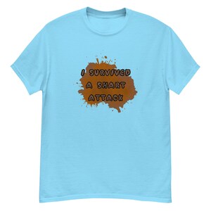 May include: Light blue t-shirt with the text "I SURVIVED A SHART ATTACK" in black letters over a brown splatter design. The shirt is a short-sleeved, crew neck style.
