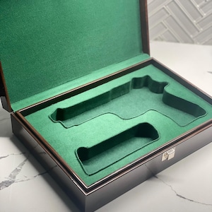 Pode incluir: Uma caixa de madeira castanho escura com um forro de veludo verde. A caixa tem um recorte para uma arma e um recorte menor para um carregador. A caixa tem uma trava prateada.