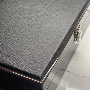 Może przedstawiać: Czarne pudełko pokryte skórą z srebrnym zatrzaskiem. Pudełko ma drewnianą ramę i fakturowaną powierzchnię.