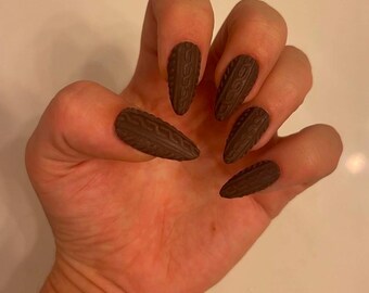 May include: Close-up of a hand with brown, long, pointed, fake nails with a knitted texture.