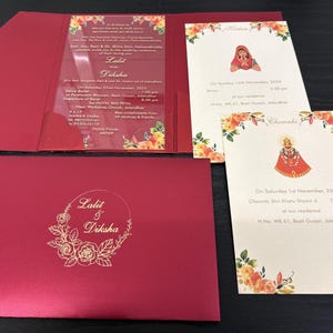 May include: Burgundy wedding invitation suite with floral accents. The suite includes an invitation, RSVP card, and a pocketfold with the names "Lalit & Diksha" in gold script. The invitation features event details.