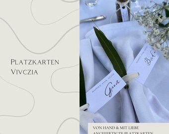 Könnte beinhalten: Weiße Platzkarte mit Kalligraphie, die "Happily Ever After" und "Groom" und "Bride" lautet, mit grünen Blättern und weißen Blumen auf einer weißen Tischdecke. Der Text "PLATZKARTEN VIVCZIA" und "VON HAND & MIT LIEBE ANGEFERTIGTE PLATZKARTEN" ist ebenfalls sichtbar.