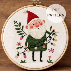May include: An embroidery hoop containing a white fabric panel with a stitched gnome wearing a red hat and green tunic, holding a string of colorful lights. The design includes green foliage and small flowers. Text reads PDF PATTERN.