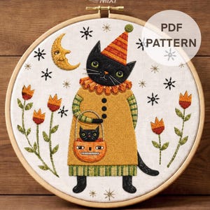 May include: An embroidery hoop containing a fabric panel with a stitched black cat wearing an orange striped hat and yellow tunic. The cat holds a pumpkin-shaped basket with a smaller cat inside. Includes a crescent moon, flowers, and stars.