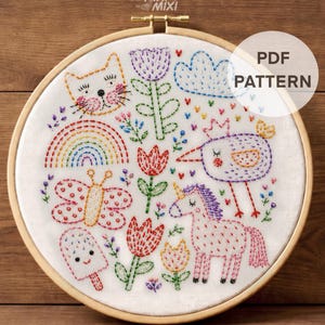 May include: A wooden embroidery hoop containing white fabric with colorful hand-stitched designs including a cat, rainbow, butterfly, popsicle, flowers, bird, and unicorn. The text reads PIXI MIXI, PDF PATTERN, and DMC Color Chart + Stitch Guide.