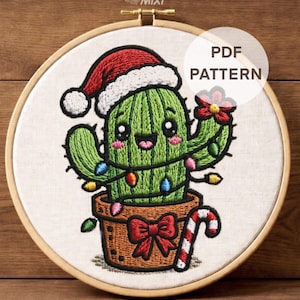 Puede incluir: Un bastidor de bordado que contiene una pieza de tela con un cactus verde bordado con un gorro de Papá Noel rojo y blanco. El cactus está decorado con luces de colores, una flor roja y se encuentra en una maceta marrón con un lazo rojo.