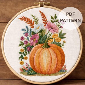 Può includere: Un telaio da ricamo contenente un pezzo di tessuto con un disegno ricamato di una grande zucca arancione circondata da fiori colorati nelle tonalità del rosa, giallo, bianco e verde. Il testo PDF PATTERN e DMC Color Chart + Stitch Guide è visibile.