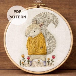 May include: An embroidery hoop containing a fabric panel with a stitched squirrel wearing a yellow cable-knit sweater and purple striped socks. The squirrel has a large, textured tail and stands among small embroidered flowers. Text reads PDF PATTERN.