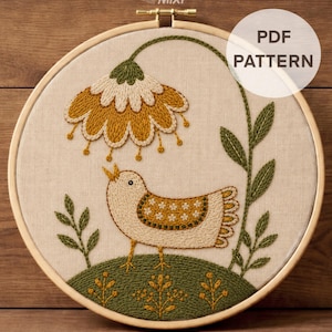 May include: An embroidery hoop containing a fabric panel with a stitched design of a bird standing on a green hill beneath a large mustard yellow flower. The bird features white and mustard yellow patterns. Text reads PDF PATTERN and DMC Color Chart + Stitch Guide.