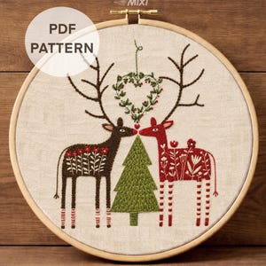 May include: An embroidery hoop containing a fabric panel with a stitched design of two deer facing a central green pine tree. The left deer is dark brown with floral patterns, and the right deer is red with floral patterns. A heart-shaped wreath hangs above.