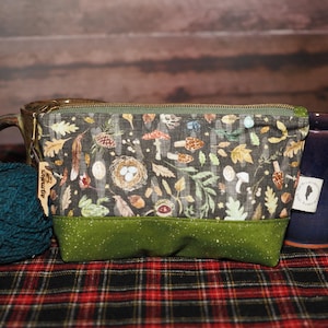 May include: A green zippered pouch with a woodland-themed fabric featuring mushrooms, leaves, and birds. The pouch has a green fabric lining and a brown leather tag that says "Sophie's".