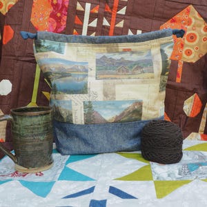May include: A drawstring project bag with a denim base and a landscape print. A ceramic mug and a ball of yarn sit beside the bag. The background features a colorful quilt.