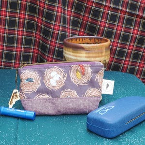 May include: A purple zippered pouch with a nest pattern, a blue glasses case, and a blue tube of lip balm are on a teal surface. A ceramic pot and a plaid fabric are in the background.