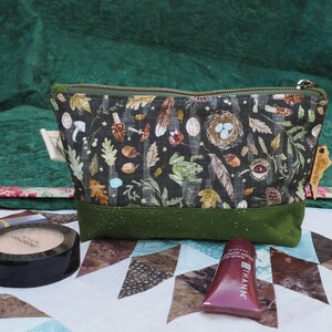 May include: A rectangular cosmetic bag with a dark background featuring a nature-themed pattern of leaves, mushrooms, and a bird's nest. The bottom is a solid green, and the zipper is green. Makeup items are in front of the bag.