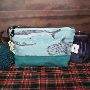 May include: A teal blue zippered pouch with a light blue background featuring a pattern of whale sharks. The pouch has a brown leather tag with the word "Seaworthy" printed on it.