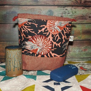 May include: A drawstring bag with a black background and a nautilus and coral design. The bag is paired with a ceramic mug and a blue glasses case. The bag is approximately 8 inches tall.