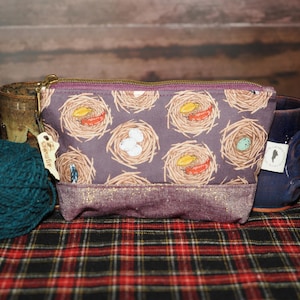 May include: A rectangular, purple zippered pouch with a nest pattern, featuring eggs and feathers. The pouch has a gold zipper and a glittery purple bottom. A ball of teal yarn and two mugs are in the background.