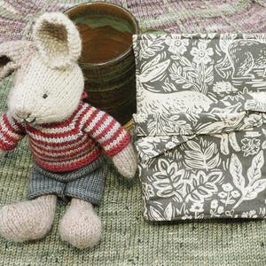 May include: A handmade knitted bunny doll wearing a red and white striped sweater and gray shorts. Beside the bunny is a gray and white floral print fabric item and a ceramic mug. The items are on a green knitted surface.