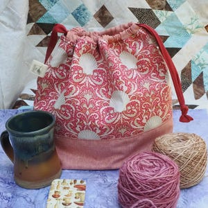 May include: A drawstring project bag featuring a red and white seashell and scroll pattern with a solid red base and red fabric ties. Beside the bag sits a two-toned blue and brown ceramic mug, a small rectangular card with mushroom illustrations, and two yarn balls.