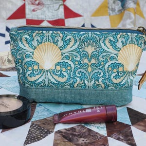 May include: A teal and cream patterned cosmetic bag with a seashell design. The bag has a teal zipper and base, and a wooden charm. A compact and a tube of makeup are in front of the bag.