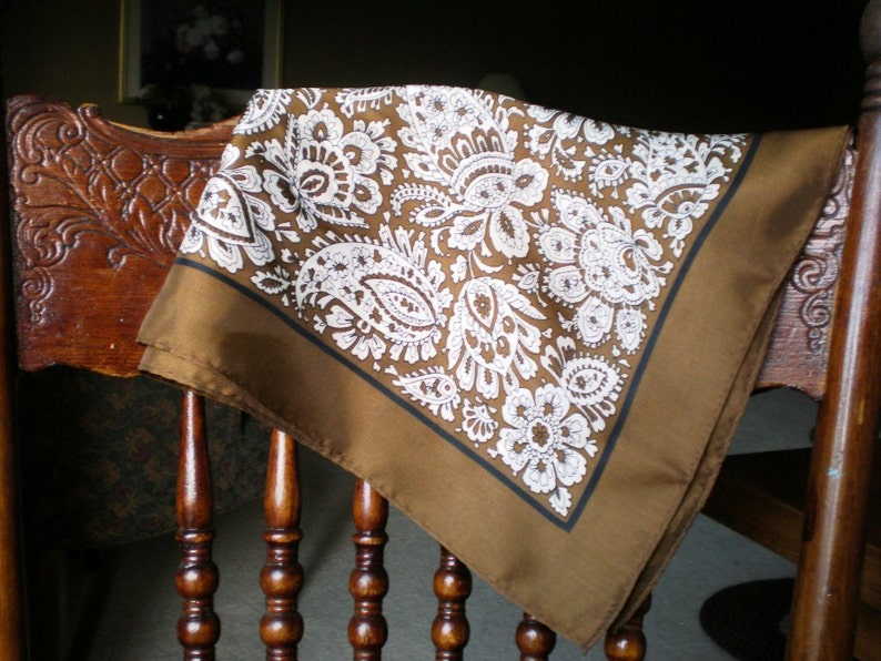May include: A brown paisley patterned bandana with white accents. The bandana is draped over a wooden chair.