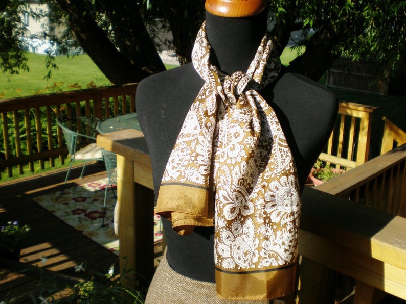 May include: A brown and white patterned scarf with a gold trim. The scarf is tied around a mannequin's neck.