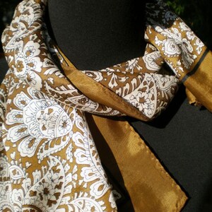 May include: A brown and white paisley patterned scarf with a gold trim. The scarf is folded and draped over a black surface.