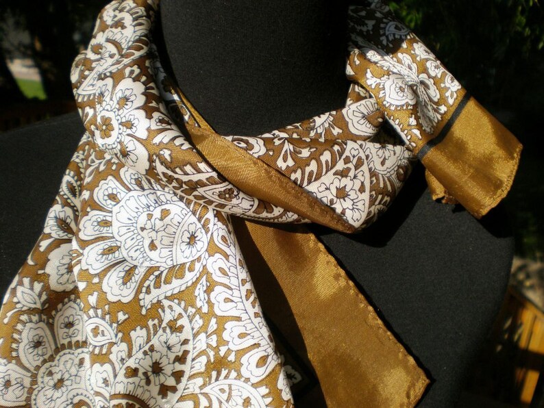 May include: A brown and white paisley patterned scarf with a gold trim. The scarf is folded and draped over a black surface.