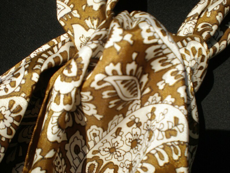 May include: A brown and white patterned silk scarf with a paisley design. The scarf is tied in a knot.