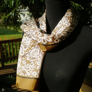 May include: A brown and white paisley patterned scarf with a gold trim. The scarf is draped around a black mannequin.
