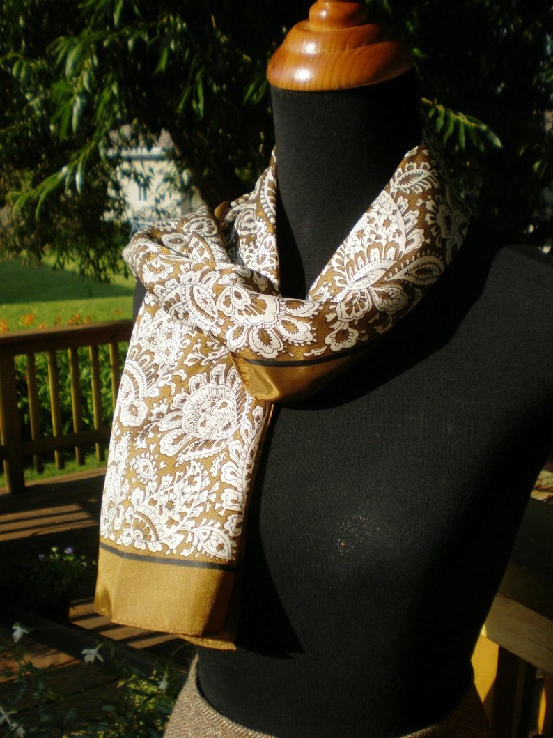 May include: A brown and white paisley patterned scarf with a gold trim. The scarf is draped around a black mannequin.