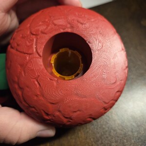 May include: A red, textured dog toy with a central opening and a yellow interior component. The toy has a spherical shape with a detailed, raised pattern across its surface. The toy is designed for pet play and engagement.