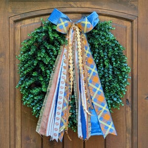 May include: A circular wreath made of dense green foliage featuring a large decorative bow at the top. The bow has long, cascading ribbon tails in various patterns, including orange pumpkin prints, blue and yellow plaid, and solid blue fabric.